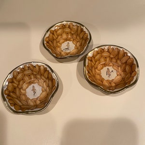 Peut inclure: Deux petits bols en céramique blanche avec un bord doré. Les bols sont remplis de formes de cacahuètes brunes et présentent un personnage de cacahuète de dessin animé au centre.