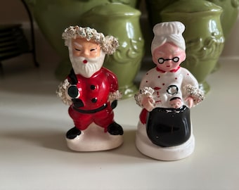 Könnte beinhalten: Ein Paar Weihnachtsfiguren aus Keramik. Einer ist der Weihnachtsmann in einem roten Anzug mit schwarzen Stiefeln, der andere ist Frau Claus in einem weißen Kleid und einer Schürze. Beide haben detaillierte Gesichtszüge und stehen auf einer weißen Oberfläche.