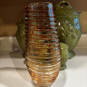 May include: A cylindrical glass vase featuring a series of horizontal, raised amber-colored glass coils wrapped around the clear body. The vase has a smooth, rounded base and an open top, showcasing a layered, ribbed texture throughout its exterior.
