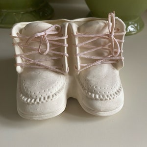 Könnte beinhalten: Ein Paar weiße Baby-Schuhe aus Keramik mit rosa Schnürsenkeln, die zu einer Schleife gebunden sind. Die Schuhe sind ein dekorativer Blumentopf.
