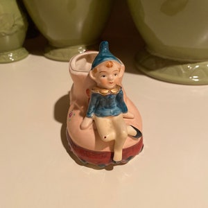 Op de afbeelding: Een keramiek beeldje van een blauwe en witte elf die op een roze schoen zit met bloemmotieven. De schoen heeft een rode en witte rand en is een plantenpot.
