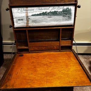 May include: A vintage wooden Chautauqua Industrial Art Desk featuring a roll-top paper display with a black and white landscape print, text reading The Chautauqua Industrial Art Desk, and a flat, light brown wooden writing surface with visible markings.