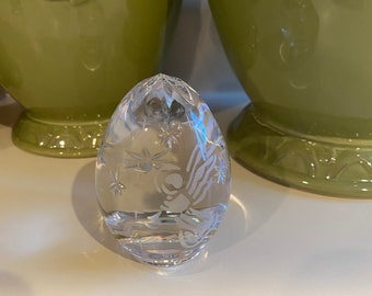 May include: A clear glass egg-shaped ornament with a delicate etched design of a snowflake and stars. The ornament is sitting on a white surface.