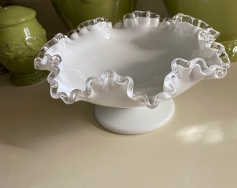 May include: A white glass bowl with a silver rim and a ruffled edge. The bowl is on a white pedestal base.