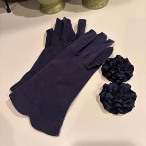 Puede incluir: Un par de guantes azul marino con borde festoneado y dos adornos de roseta azul marino a juego. Los guantes son de tela suave, probablemente para eventos formales u ocasiones especiales.
