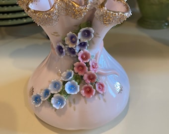 May include: A pink porcelain vase with a ruffled top and gold trim. The vase is decorated with 3D flowers in shades of pink, blue, and white.
