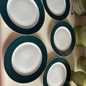 Puede incluir: Una colección de platos ovalados blancos con un borde verde oscuro. Los platos varían en tamaño y están dispuestos sobre una superficie beige claro. Se ve vajilla adicional en tonos verdes en el fondo.