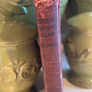 May include: Close-up of the spine of a vintage book titled "Stevenson's Treasure Island" with the author's name "Gaston" visible. The book's cover is a faded burgundy color, showing signs of wear. The background features green ceramic vases.