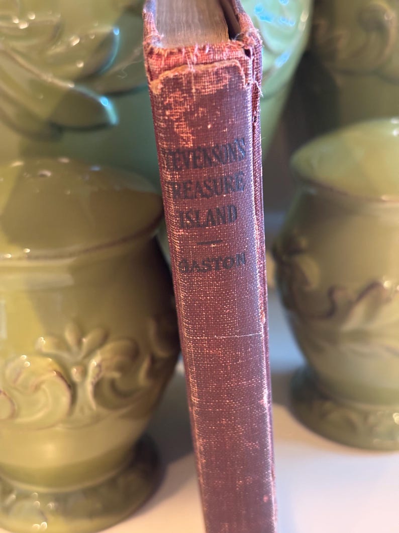 May include: Close-up of the spine of a vintage book titled "Stevenson's Treasure Island" with the author's name "Gaston" visible. The book's cover is a faded burgundy color, showing signs of wear. The background features green ceramic vases.