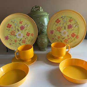 May include: A collection of yellow and floral-decorated dinnerware. The set includes plates, cups, saucers, and bowls. A green ceramic jar and a small container are also present. The plates feature a yellow rim and a floral design with red, orange, and white flowers.