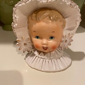 May include: A white ceramic figurine of a baby girl wearing a bonnet and a ruffled collar. The figurine has blue eyes, rosy cheeks, and a sweet smile. The bonnet and collar are decorated with gold trim.