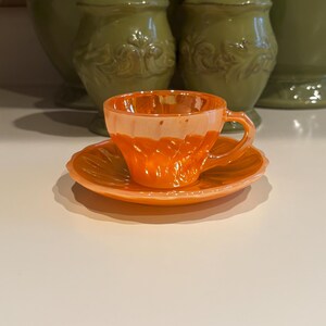 May include: A vibrant orange iridescent glass teacup and saucer set. The teacup features a ribbed, fluted design with a matching handle, while the saucer has a scalloped edge with a radiating petal pattern. The glass has a glossy, reflective finish.