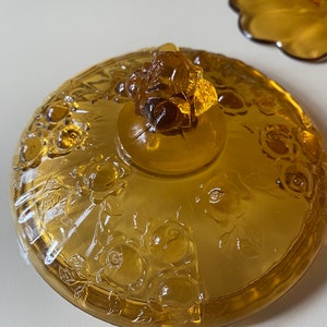 K&ouml;nnte beinhalten: Amberfarbener Glasdeckel mit Rosenmuster und einem Rosenknospenknopf. Der Deckel ist mit einem floralen Muster gepr&auml;gt.