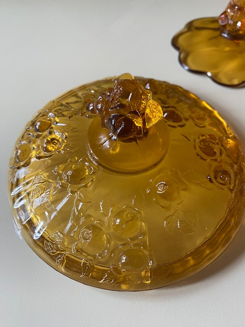 K&ouml;nnte beinhalten: Amberfarbener Glasdeckel mit Rosenmuster und einem Rosenknospenknopf. Der Deckel ist mit einem floralen Muster gepr&auml;gt.