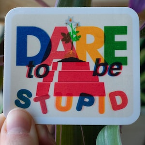 May include: A square sticker with the words "DARE to be STUPID" in colorful block letters. The sticker has a white background and a small illustration of a plant. The sticker is held in front of a plant.