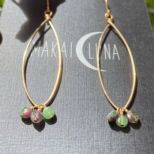 May include: Gold oval hoop earrings with three green and pink gemstone drops. The earrings are on a black background with the text "Makai Luna" visible.