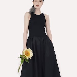 May include: A woman wearing a black sleeveless dress with a fitted bodice and a flowing skirt. She is holding a single sunflower.