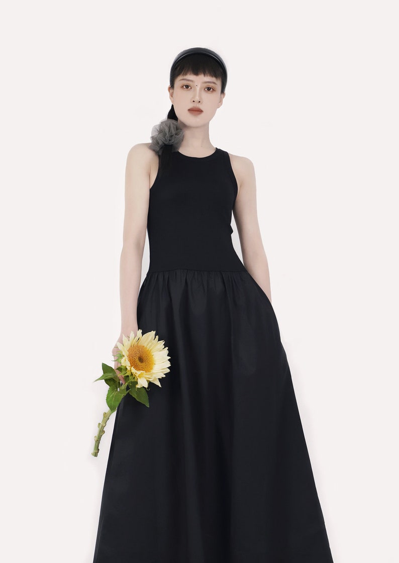 May include: A woman wearing a black sleeveless dress with a fitted bodice and a flowing skirt. She is holding a single sunflower.