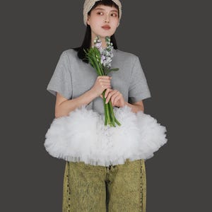 May include: A woman wearing a grey t-shirt, a white tulle skirt, and light green jeans. She is holding a bouquet of flowers and wearing a beige knitted headband.