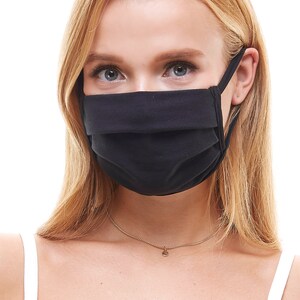 May include: A black face mask with pleats, worn by a person with long blonde hair. The mask has thin straps that go around the ears.
