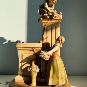 Op de afbeelding: Een porseleinen beeldje van twee mensen, een vrouw die een mand met bloemen vasthoudt en een man die naar bloemen reikt op een pilaar. Het beeldje is geschilderd in tinten bruin, groen en wit.