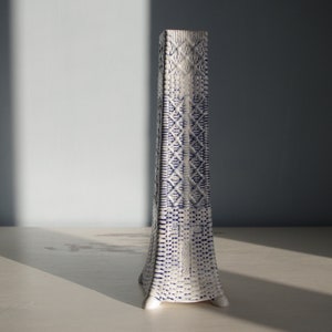 Könnte beinhalten: Eine hohe, weiße Keramikvase mit einem blauen und weißen geometrischen Muster. Die Vase hat einen quadratischen Boden und einen schmalen Hals.