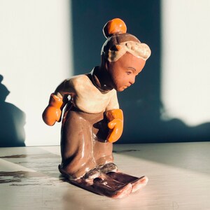 May include: A ceramic figurine of a person wearing a brown hat and orange mittens, skiing down a slope. The figurine is painted in shades of brown, orange, and white.
