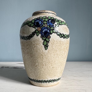 Op de afbeelding: Een beige keramische vaas met een getextureerd oppervlak. De vaas heeft een decoratief bloemmotief met blauwe en groene accenten. Het ontwerp is in een guirlande patroon rond de vaas gerangschikt.