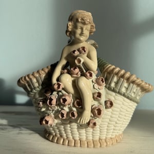 May include: A ceramic figurine of a cherub seated in a woven basket, holding flowers. The cherub is light-skinned with curly hair, and the basket is cream-coloured with pink and brown accents. The basket is filled with pink roses.