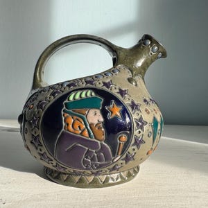 May include: A ceramic pitcher with a handle and spout, decorated with a figure in a turban within a medallion. The design includes stars and geometric patterns in beige, blue, green, and orange. The pitcher is a decorative item.