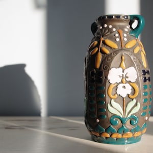 May include: A decorative ceramic vase with a dark brown base colour and colourful floral and geometric patterns. The vase has a teal rim and handles, with white, yellow, and blue accents. The vase is on a white surface.