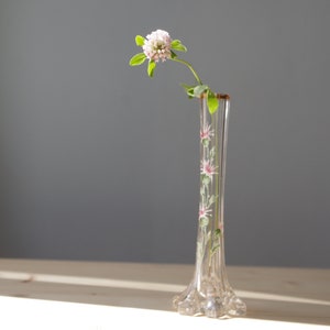 Puede incluir: Un jarrón de cristal transparente con base acampanada y cuello esbelto, adornado con diseños florales rosas y blancos pintados a mano. Una sola ramita de una flor de trébol rosa con hojas verdes se coloca dentro del jarrón. El jarrón mide unos 20 cm de altura.