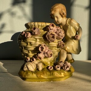 May include: A ceramic figurine of a child in a yellow dress, next to a basket-shaped vase. The vase is decorated with pink flowers and sits on a green base. The figurine is likely vintage and is a decorative item.