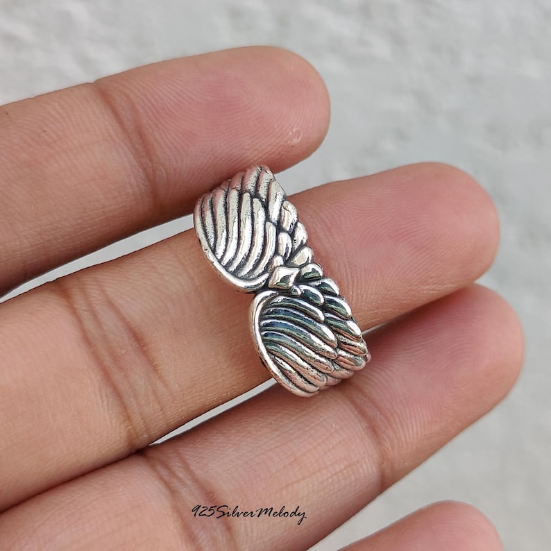 May include: A silver ring featuring a detailed design of two symmetrical wings meeting in the center. The metal has an oxidized finish to highlight the carved feather textures. The text 925 Silver Melody is printed at the bottom of the image.