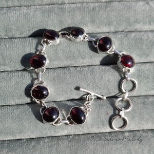 May include: A silver bracelet with a toggle clasp features a chain of oval-shaped red gemstones.