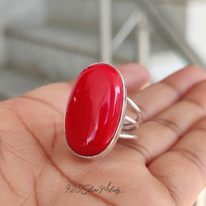 May include: A striking red gemstone ring with a silver band. The oval-shaped, polished red stone is the focal point, set in a minimalist silver design. The ring is held in a hand. The text "925 Silver Melody" is visible.