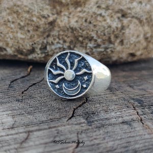 May include: A silver signet ring featuring a circular face with a dark, textured background. The design depicts a raised sun with radiating rays positioned above a crescent moon and two small four-pointed stars. The text 925 Silver Melody is visible.