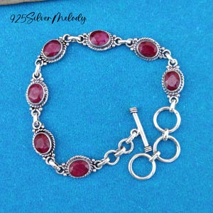 May include: A silver bracelet with a toggle clasp features oval-shaped red gemstones. The bracelet is on a blue background.