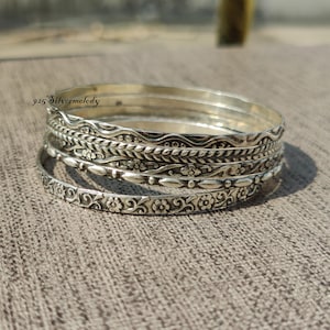 Peut inclure: Ensemble de quatre bracelets en argent avec des motifs floraux et géométriques complexes. Les bracelets sont empilés les uns sur les autres, mettant en valeur les différents motifs. Le texte "925 Silvermelody" est visible sur le bracelet du haut.