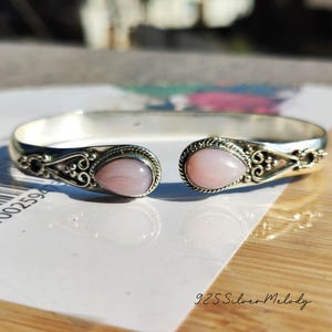 May include: A silver bangle bracelet with two pink oval-shaped stones. The bracelet features a delicate floral design.