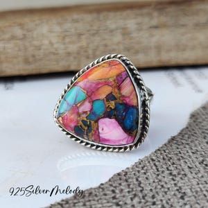 May include: A triangular silver ring with a colorful, multi-faceted gemstone set in a rope-like border.