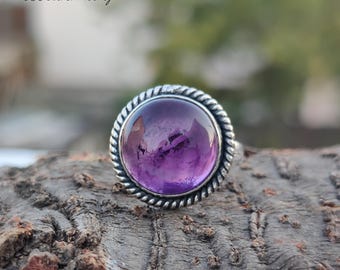 May include: A silver ring with a round, purple amethyst gemstone. The ring has a rope-like design around the edge of the setting.