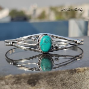 Puede incluir: Un brazalete de plata con una piedra turquesa engastada en un óvalo de plata. El brazalete tiene un diseño delicado y ornamentado con espirales y pequeñas cuentas.