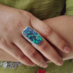 May include: A silver ring featuring a large, rectangular, iridescent green opal stone. The ring is set in a silver band with a beaded edge.