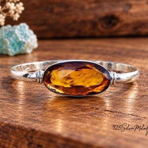 May include: A sterling silver bangle bracelet featuring a large, oval-shaped, faceted amber-coloured gemstone set in the centre. The metal band has a smooth, polished finish with decorative ribbed detailing on either side of the stone setting.