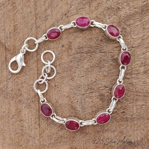 May include: A silver bracelet with oval-shaped, deep red gemstones. The bracelet features a lobster clasp closure and is set against a wooden background. The gemstones are evenly spaced along the silver chain.