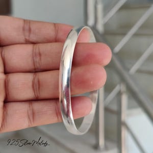 May include: A silver bangle bracelet with a polished finish. The bracelet is shown on a person's hand.