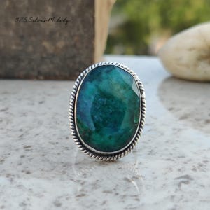 Peut inclure: Une bague en argent avec une grande pierre précieuse ovale de couleur vert foncé. La bague a une bande en argent torsadée décorative.