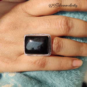 May include: A silver ring with a black rectangular gemstone. The ring is on a finger.