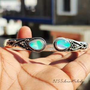May include: A silver bangle bracelet with two iridescent opal stones set in the center of each side. The bracelet is decorated with intricate silver filigree.